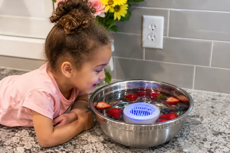 Child watching PuriBreeze produce cleaner in a bowl with fresh fruit in a family kitchen
